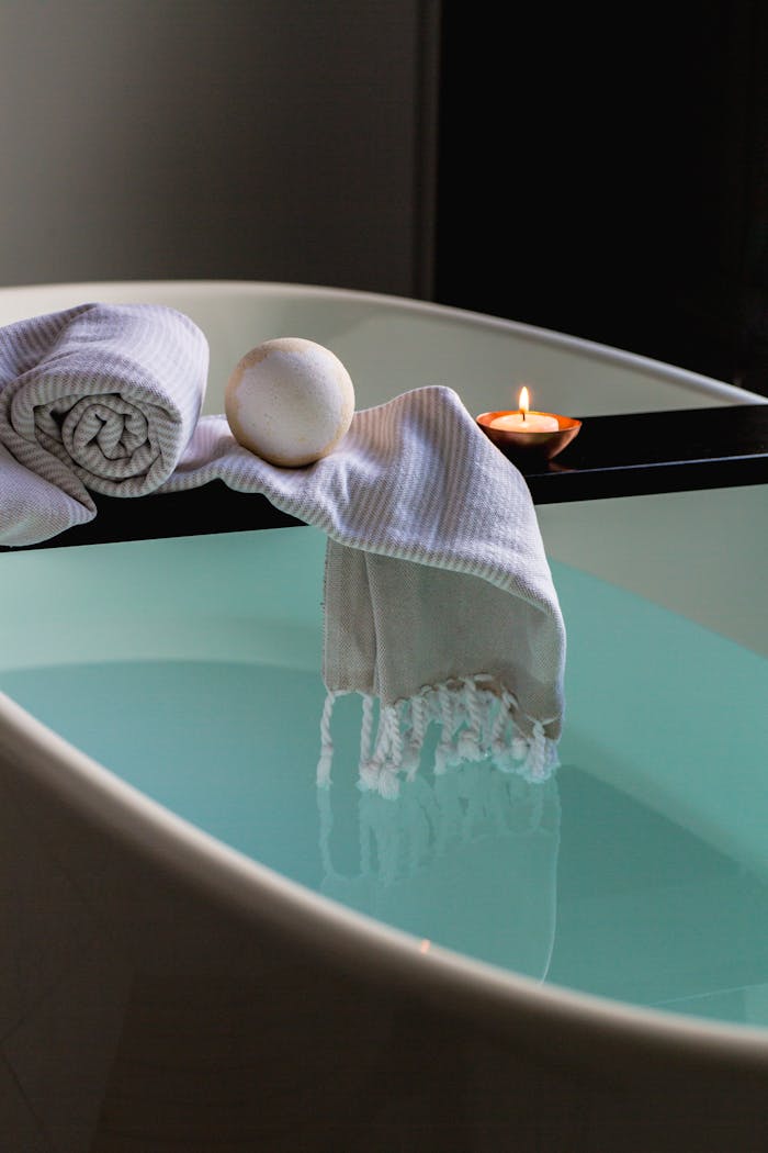 Elegant indoor spa setting with bathtub, candle, towel, and bath bomb for relaxation.